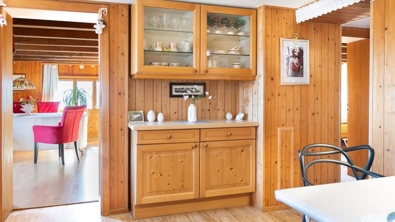 light wooden cupboard, cabinets, shelves, plate on table, framed photos, chairs, flower vase, table and red chairs in another room, glass, ceramic ware, and plates