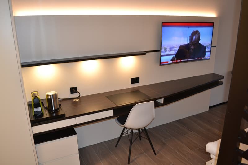 The image shows a modern home office setup with a large desk, a mounted TV, and various decor items like a wine bottle and a canister. The room has a minimalist and sleek design with white walls, dark wood flooring, and recessed lighting.