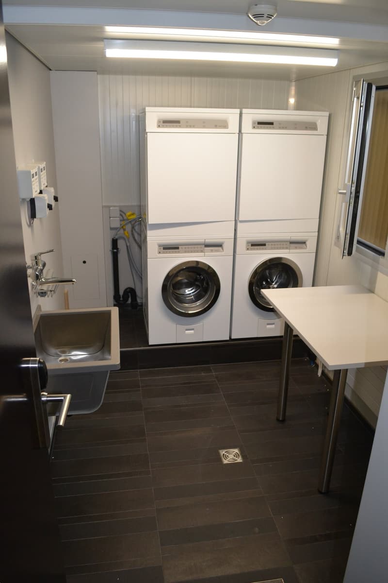 The image shows a laundry room with two stacked washing machines, a dishwasher, and a utility sink. The room has white walls, a tiled floor, and overhead lighting fixtures.
