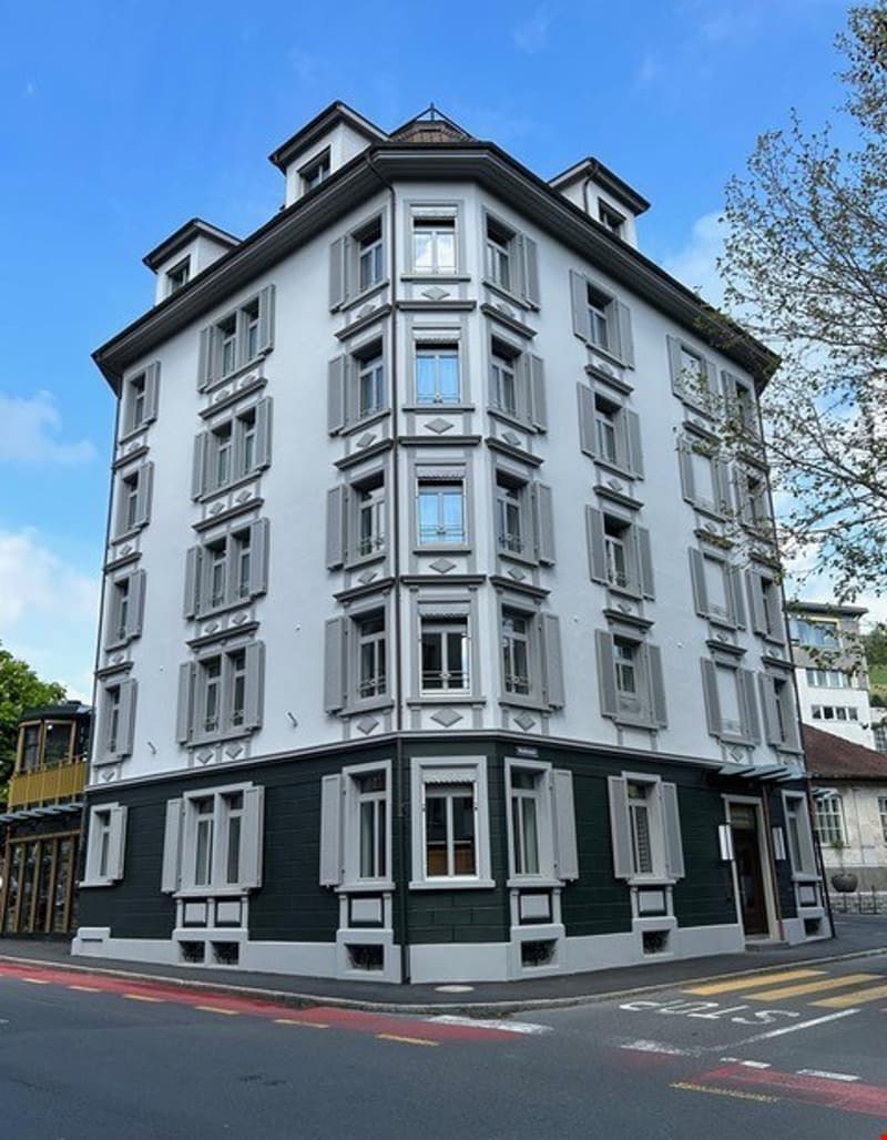 The building has a contemporary architectural design with white and green trims, featuring multiple stories with numerous windows. The building is located on a corner with a road and street light nearby.