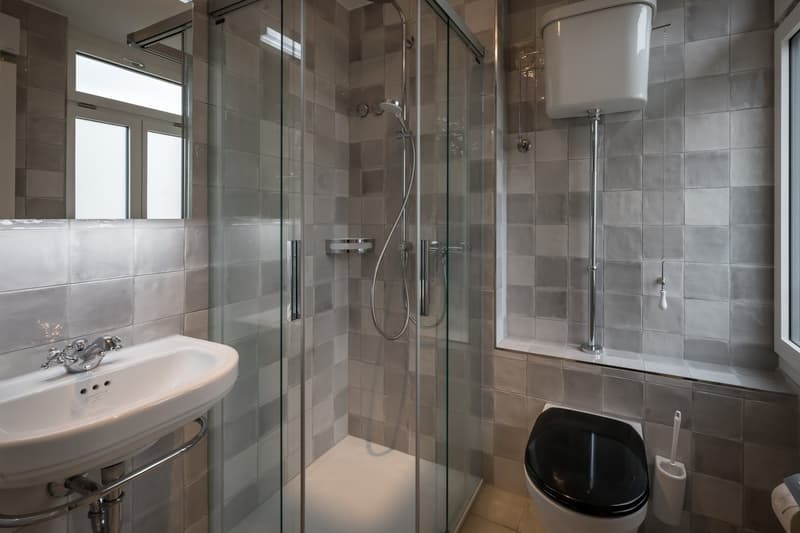 The image shows a modern bathroom with a glass-enclosed shower, a pedestal sink, and a black toilet. The bathroom is tiled with gray and beige tiles, and there is a large window that provides natural light. The bathroom appears to be well-designed and equ