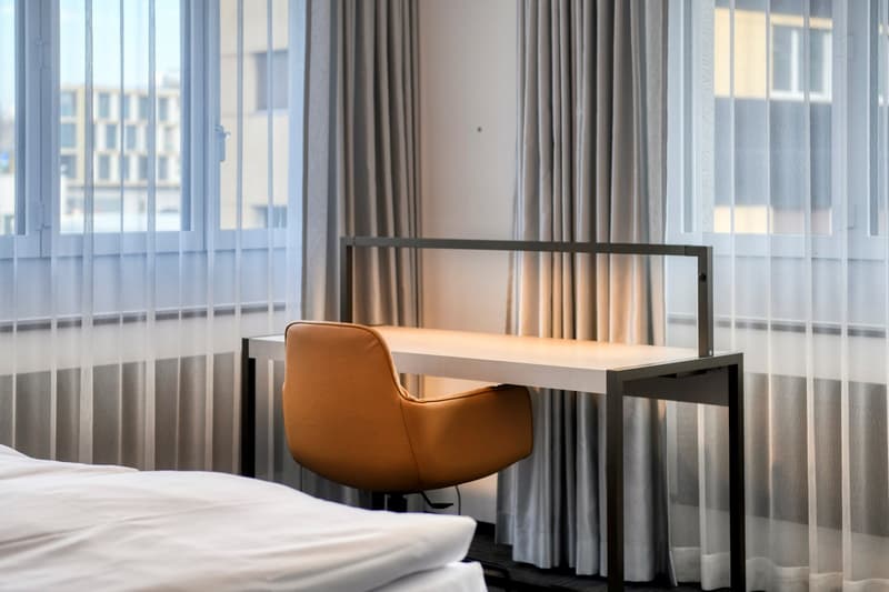 Spacious bedroom with large windows, gray curtains, a modern desk and chair, and a comfortable-looking bed.