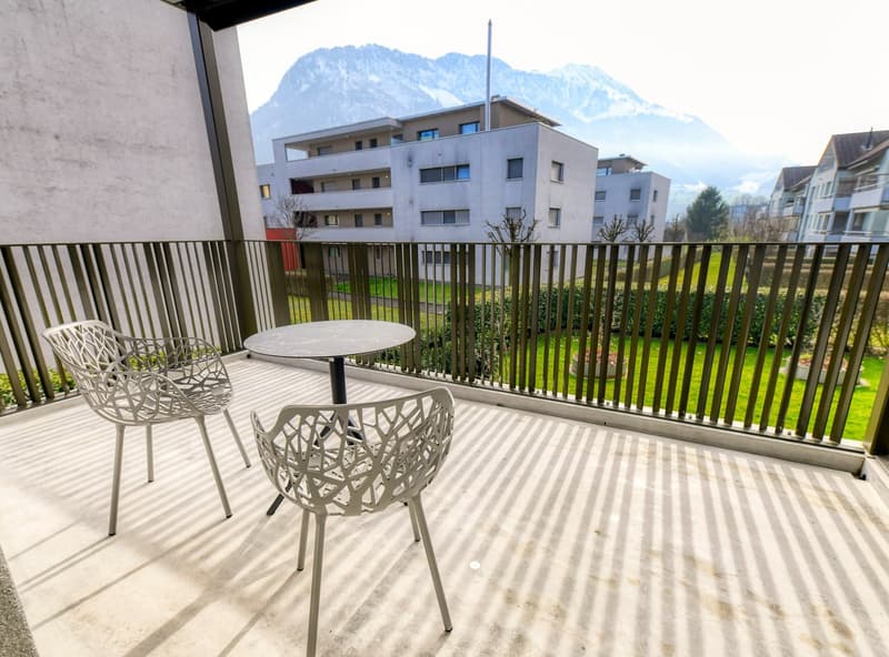 The image shows a balcony or terrace of an apartment building. The balcony has a table and two chairs, and overlooks a grassy area with trees and other apartment buildings in the background. The building appears to be a modern, multi-story structure with