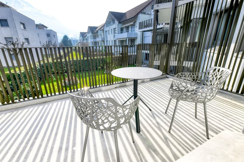 Balcony with white patio furniture, including a round table and two chairs with intricate lattice-style designs. The balcony overlooks a grassy area and other apartment buildings.