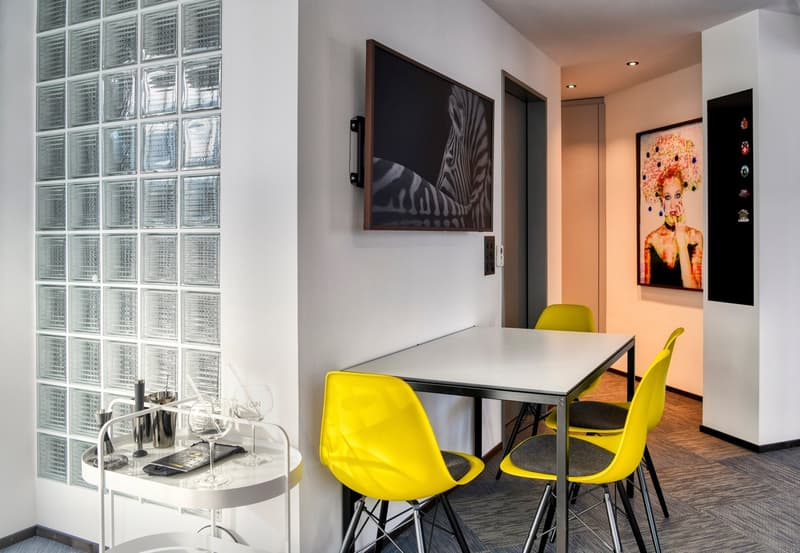 Modern apartment with glass block wall, dining table with yellow chairs, artwork on the wall, and a bar area with wine glasses and a TV.