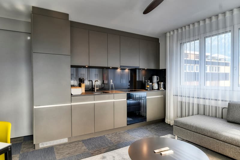 The image shows a modern studio apartment with a fully equipped kitchen. The kitchen features sleek gray cabinets, a built-in dishwasher, and stainless steel appliances. The living area has a gray sofa and a coffee table. Large windows provide natural lig