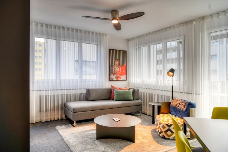 Spacious living room with large windows, gray sofa, coffee table, colorful accent pillows, and a ceiling fan. The room has a modern and cozy feel with the neutral color scheme and pops of color from the artwork and decor.