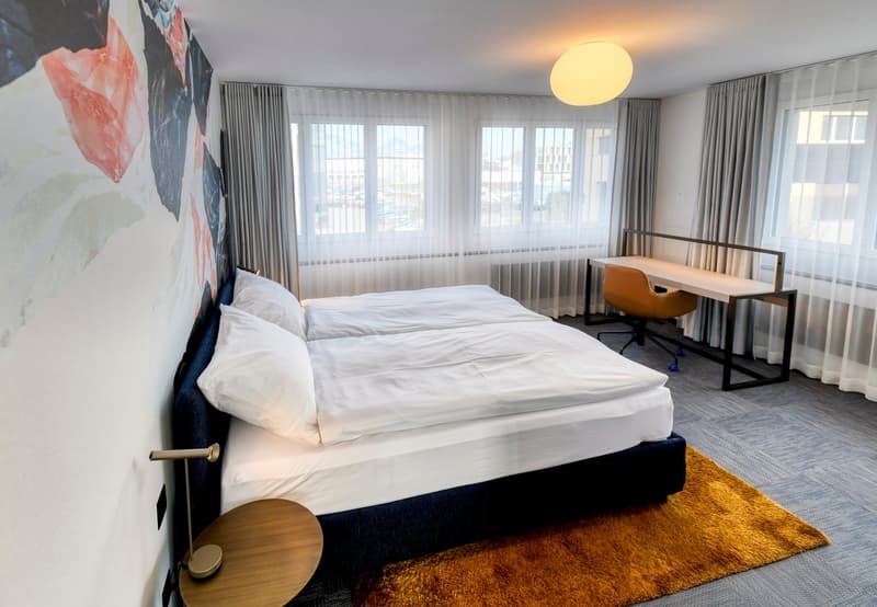 Spacious bedroom with large windows, white bedding, modern furniture including a desk and chair, and a colorful abstract art piece on the wall.