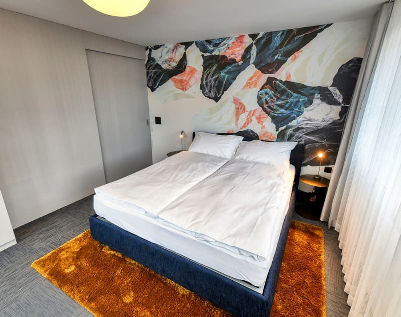 Spacious bedroom with a large abstract mural wall, a king-size bed with white bedding, a navy blue bed frame, and an orange shag rug on the floor. The room also has a window with curtains and a small side table with a lamp.