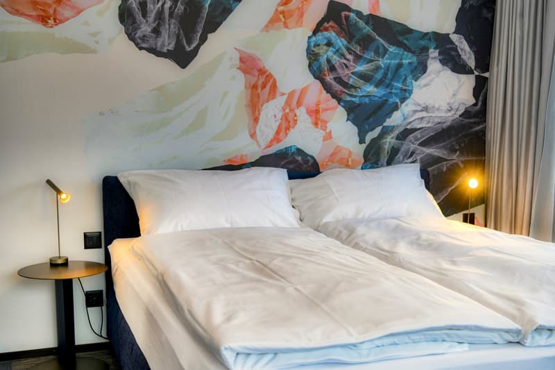 Bedroom with a large floral wallpaper, white bedding, and a side table with a lamp