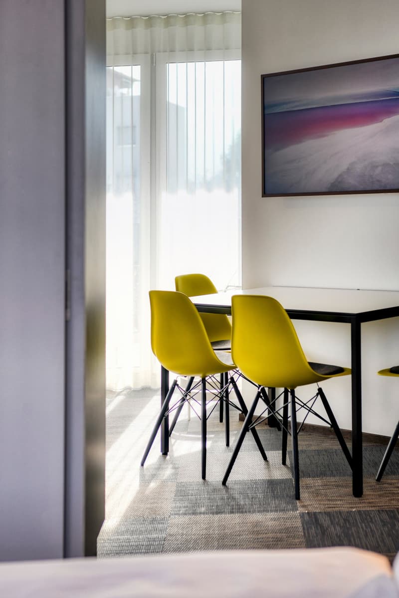 The image shows a modern dining room with a black rectangular table and four yellow chairs. The room has large windows that provide natural light and a view of what appears to be a balcony or terrace. The walls are a neutral color, and there is a framed a
