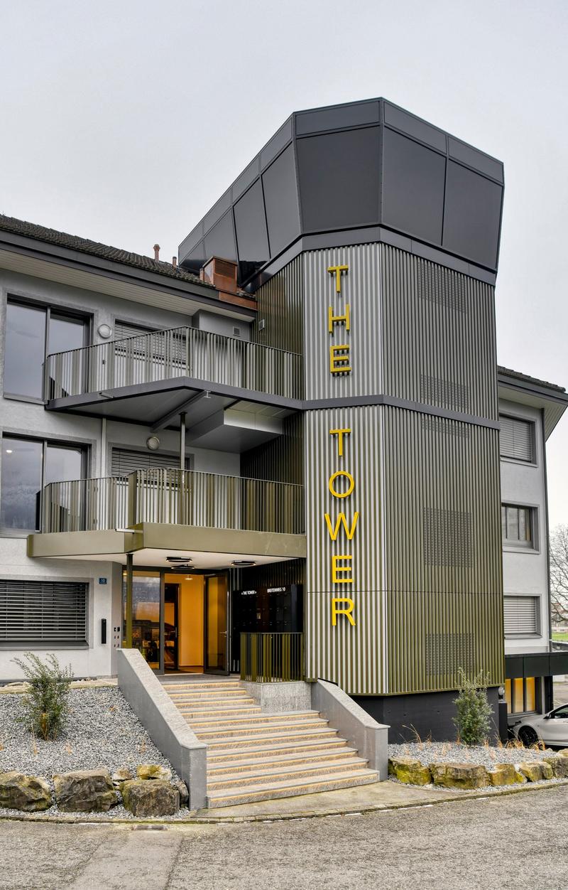 Modern multi-story building with a distinctive black and yellow facade, featuring balconies on the upper floors and a prominent entrance with the word 'Tower' displayed in large yellow letters.