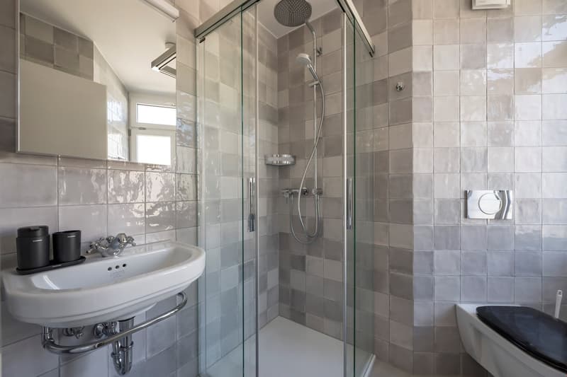 Spacious bathroom with gray tile walls, glass shower enclosure, pedestal sink, and modern fixtures