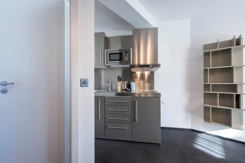 Compact kitchenette with stainless steel appliances, including a microwave and dishwasher, built-in cabinets, and a sink with a faucet. The space also includes a shelving unit for storage.
