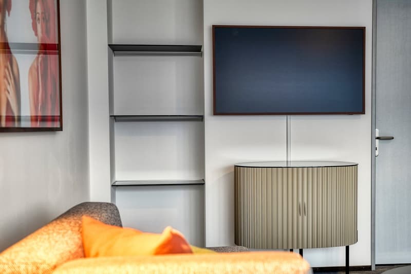 Modern living room with a large TV mounted on the wall, a shelving unit, and a lamp with a pleated shade. The room also features a couch with orange and gray pillows, suggesting a cozy and comfortable space.