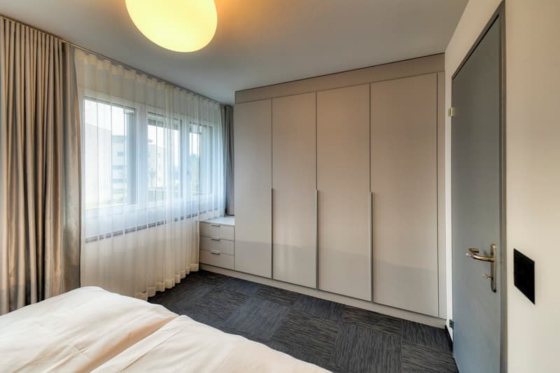 Spacious bedroom with large windows, beige walls, built-in wardrobe, and dark gray carpeted floor