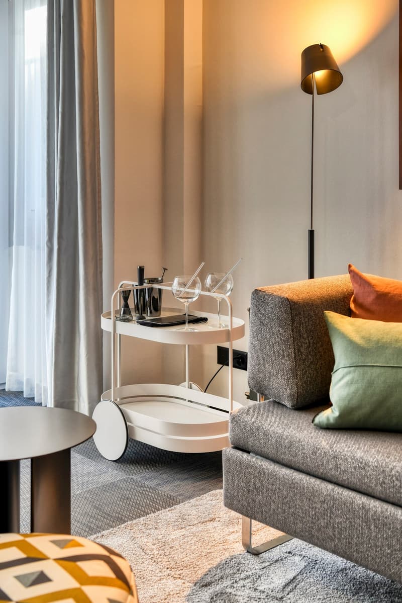Cozy living room with gray sofa, decorative pillows, white side table with wine glasses and bar accessories, floor lamp, and curtains covering a window or balcony door