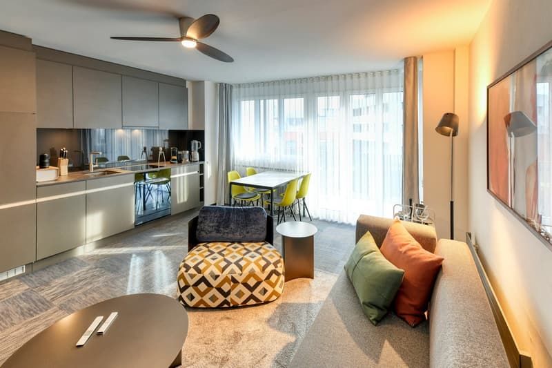 Open-concept living room and kitchen with large windows, modern gray cabinets, stainless steel appliances, dining table with yellow chairs, patterned ottoman, and couch with colorful pillows.