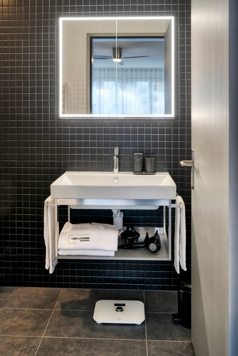 Bathroom with black tile walls, white sink vanity, lighted mirror, and a dishwasher visible on the counter