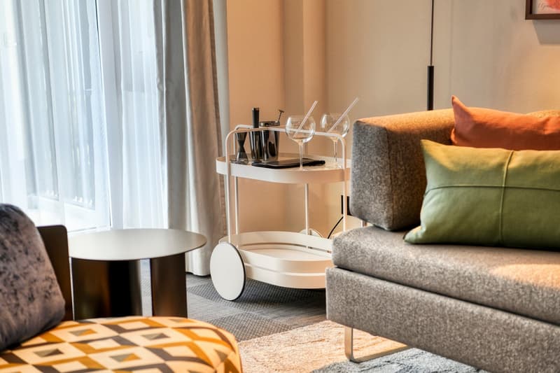 Cozy living room with large windows, gray sofa, colorful throw pillows, coffee table, and a bar cart with wine glasses and bottles.