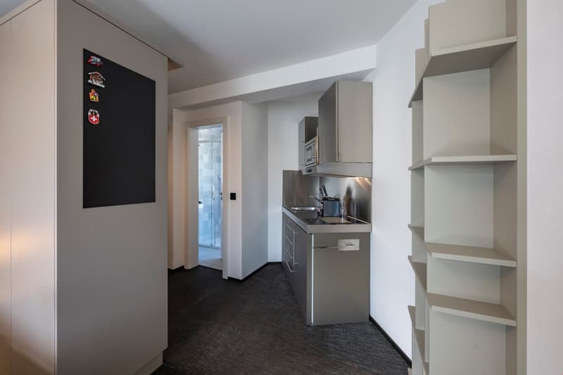 Compact studio apartment with open kitchen, stainless steel appliances including a dishwasher, shelving units, and a blackboard wall for notes or decoration.