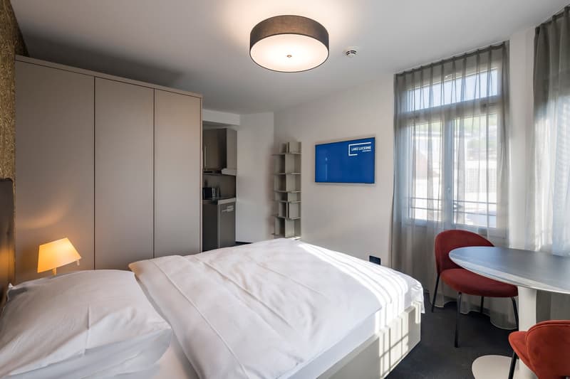 Bedroom with a double bed, white linens, side table with lamp, a built-in wardrobe, small kitchenette, and a flat-screen TV.