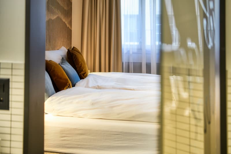 Cozy bedroom with a large bed, white bedding, and brown and blue accent pillows. The room has large windows with sheer curtains, providing a bright and airy atmosphere. There appears to be a balcony or terrace visible through the windows.