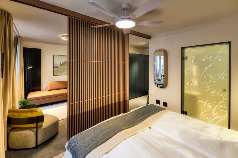 Spacious bedroom with wooden slat divider, large windows, modern furniture including a sofa, and a decorative wall art piece