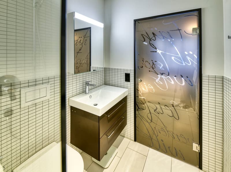 Modern bathroom with white tile walls, vanity with sink, and a frosted glass door with abstract artwork.