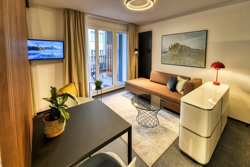 Spacious living room with large windows, balcony access, modern furniture including a sofa, coffee table, and side tables, wall-mounted TV, and decorative plants.