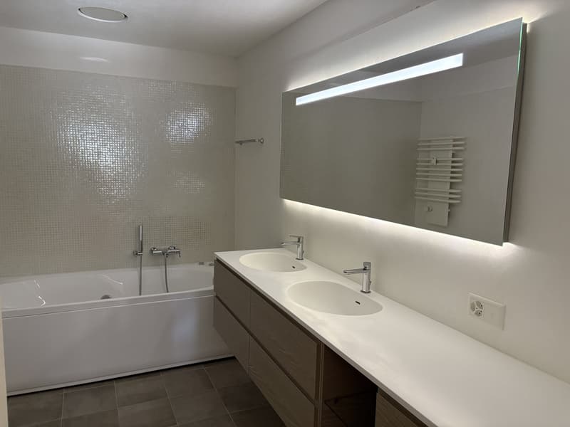 modern bathroom with white bathtub, two sinks with faucets, mirror with built-in light, towel heater, tiled flooring