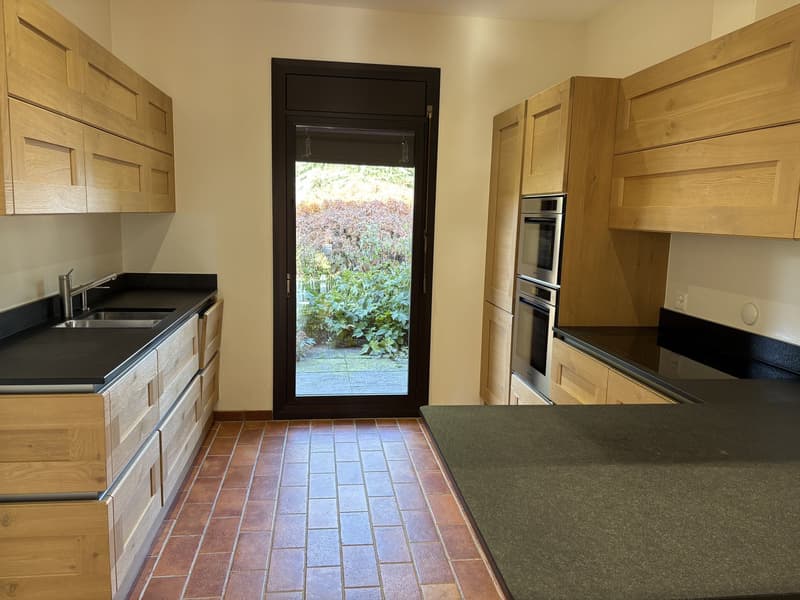 Modern open-plan kitchen with island, brick flooring, double oven, wooden cabinetry, black countertops, glass door to outdoor area.