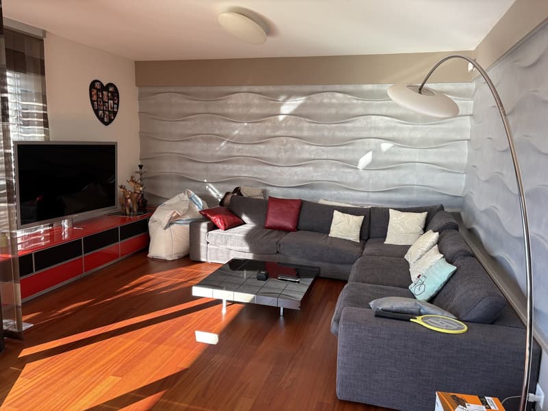 Modern living room with gray couch, red pillows, wooden floor, white ceiling lamp, coffee table, large screen TV, floor lamp, white decorative wall
