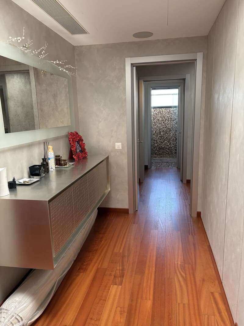 Hallway with shiny wooden flooring, walls are metallic and white, large mirror, desk with bottle, mirror and bowl on top, hallway leading to other rooms