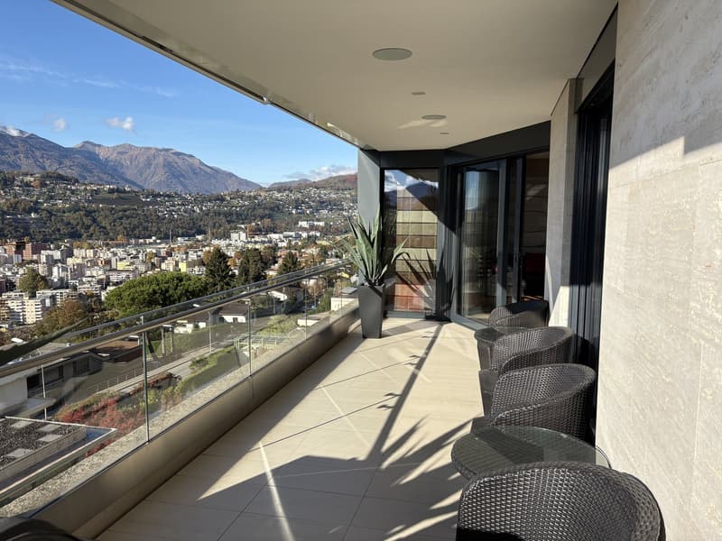 Spacious balcony with glass railings, tiled floor, outdoor seating, and a view of the city and mountains