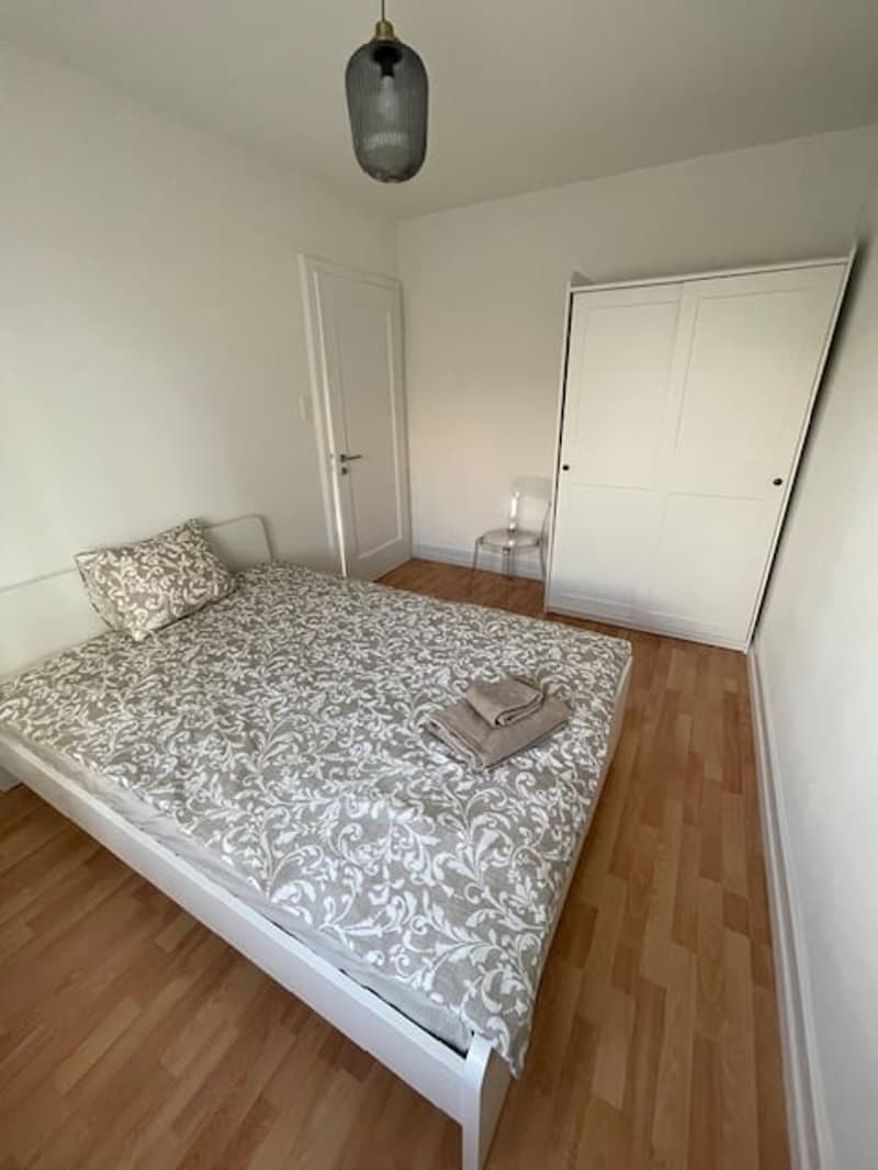 white walls, wooden floor, a double bed with bedspread, a white wardrobe, and a white door, and a chair