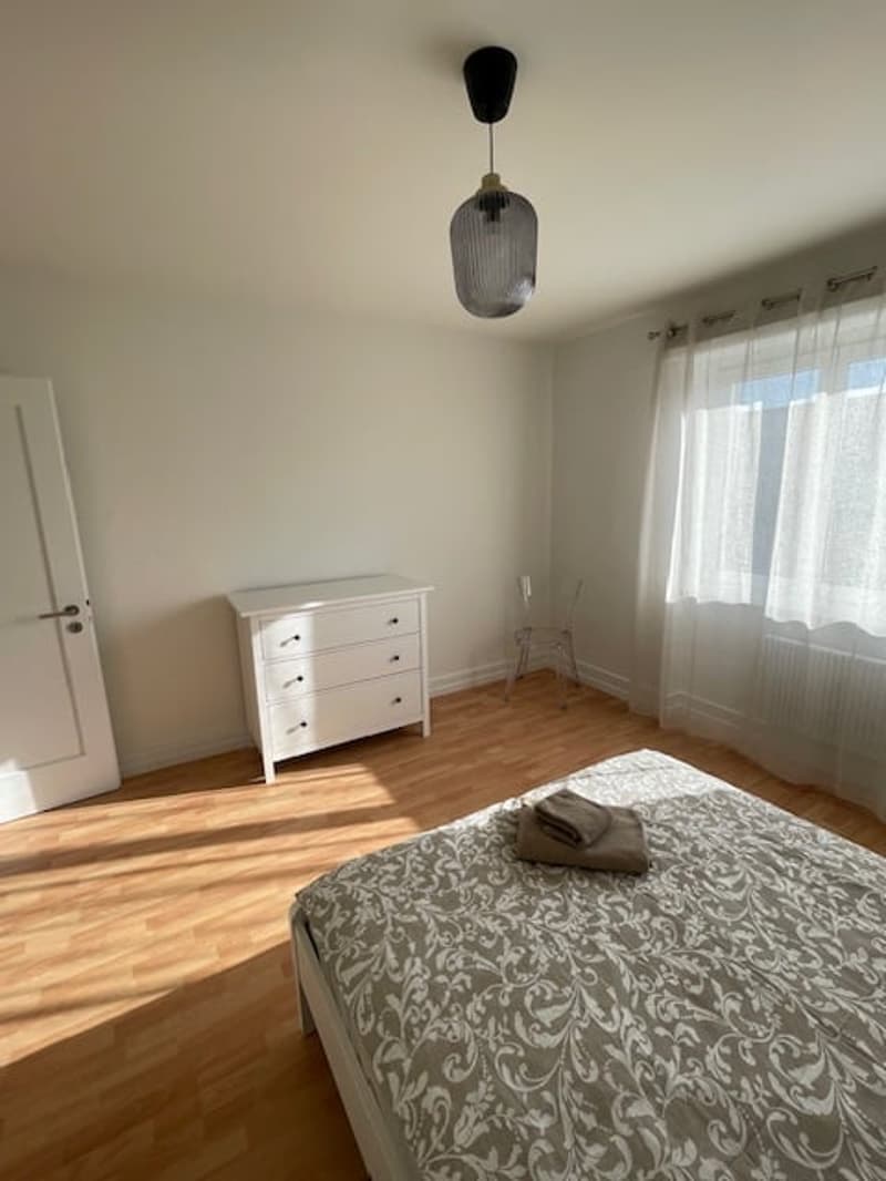 Bedroom with bed, white walls, wooden floor, white chest of drawers, white door, window with white curtains, a lamp hanging from the ceiling, and a chair
