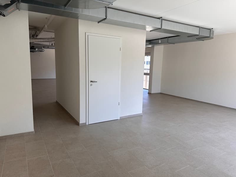 An empty room with tiled flooring, white walls, and a door, possibly used for storage or waiting. There are visible ceiling pipes and lights.