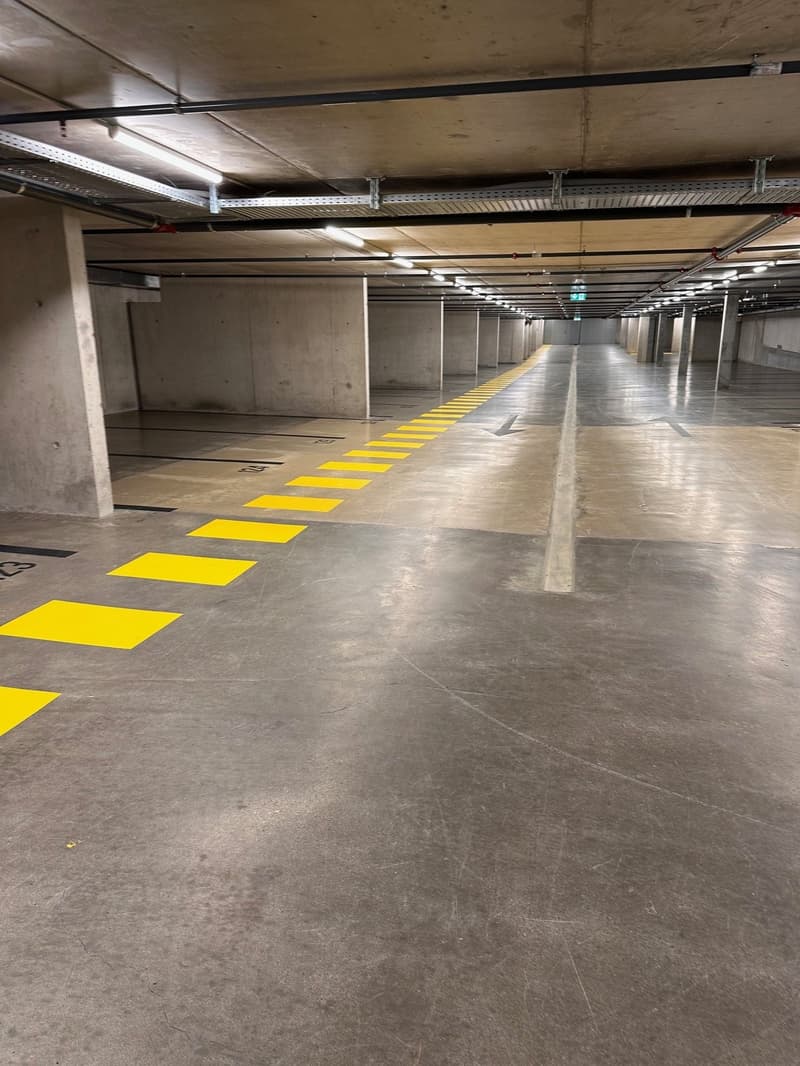 An indoor parking area with concrete floors, yellow painted parking slots, white ceiling lights, and yellow directional arrows.