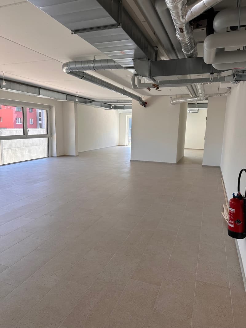Large room, tiled flooring, industrial pipework, white walls, and large window.