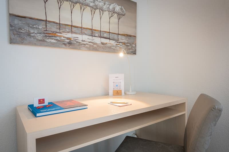 Desk with books, laptop, and WiFi sign, wall art depicting a landscape, lamp, and comfortable chair