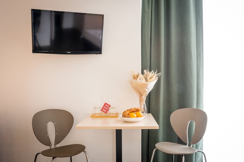 The image shows a hotel room with a flat-screen TV mounted on the wall, a small table with a bouquet of dried flowers, a plate of croissants and fruit, and two gray chairs. There appears to be a kitchenette area with a dishwasher visible.