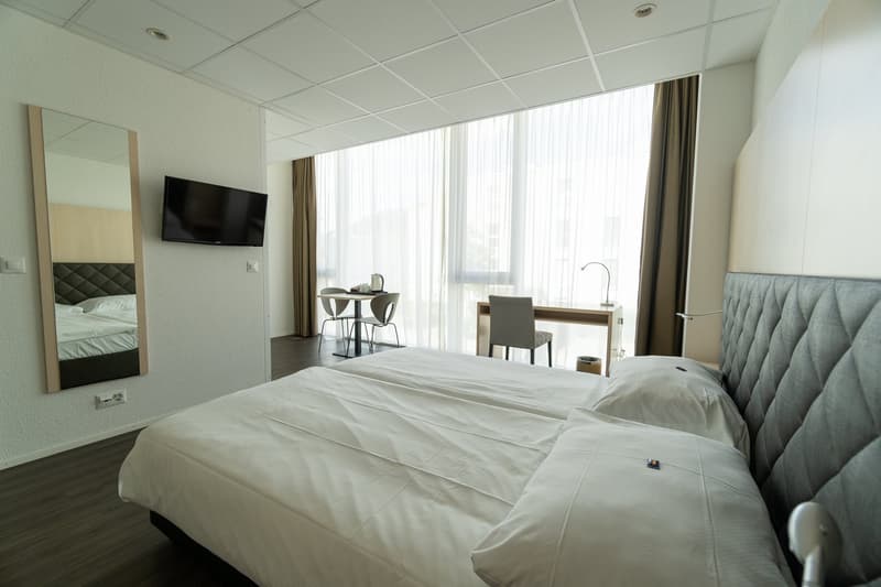 Spacious hotel room with large windows, a king-size bed with a tufted headboard, a desk and chair, and a flat-screen TV mounted on the wall.