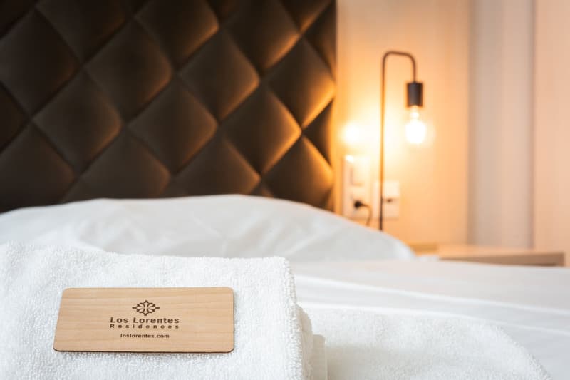 Cozy bedroom with textured wall panels, a white bedsheet, and a wooden sign with the logo of 'Los Lorentes Residences'. A lamp is visible, suggesting a warm and inviting atmosphere.