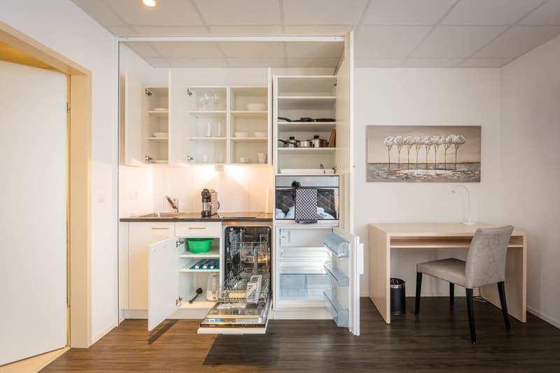 Compact kitchen with white cabinets, open shelving, stainless steel appliances including a dishwasher, and a small dining area with a table and chair