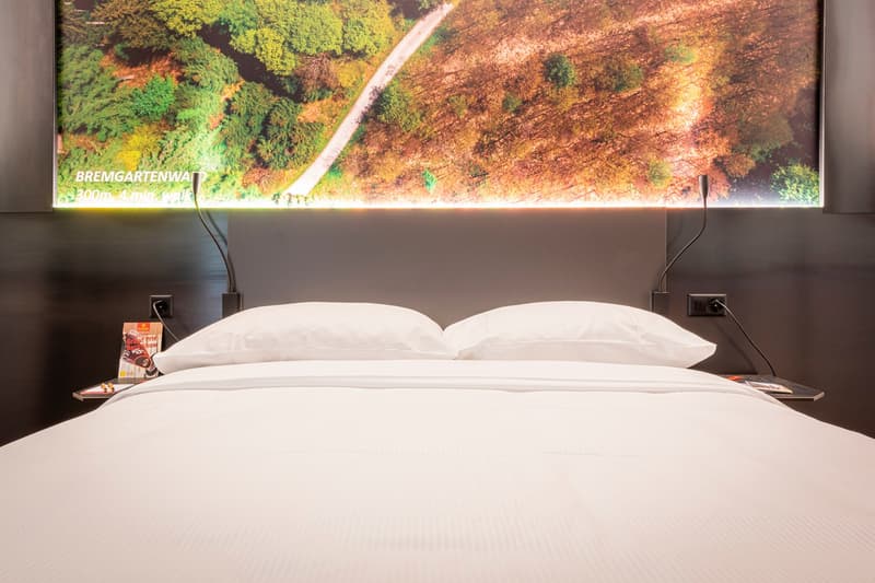 The image shows a hotel room with a large bed and a scenic landscape displayed on a screen or monitor behind the bed. The room has a minimalist and modern design, with white bedding and a simple nightstand on each side of the bed.