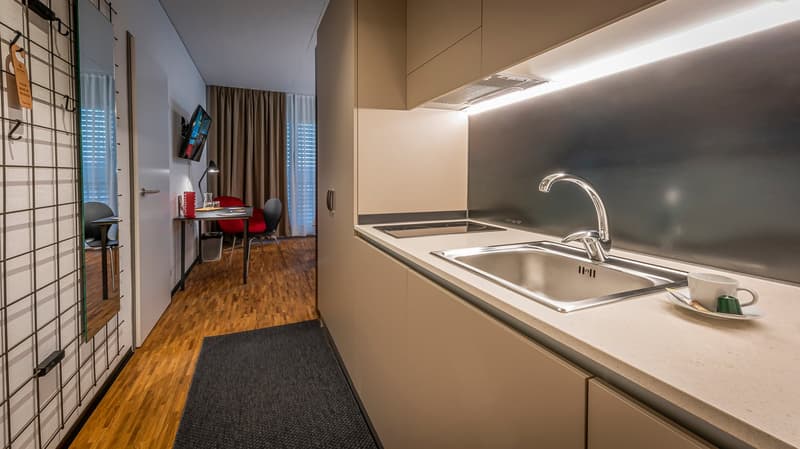 The image shows a studio apartment with a kitchenette area featuring a sink, countertop, and what appears to be a dishwasher. The living space has a desk and chair, and the flooring is hardwood. The room has large windows and curtains, providing natural l