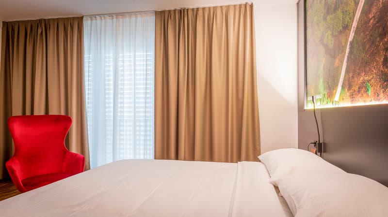 Spacious bedroom with large windows, beige curtains, a red armchair, and a white bed with bedding.