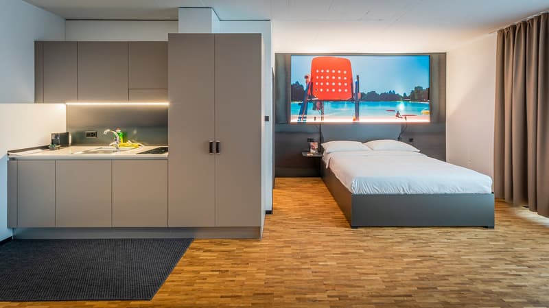 small room with a bed, a kitchen area, and a large screen