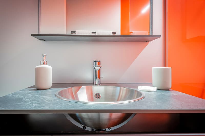 Modern bathroom with stainless steel sink, soap dispenser, and white ceramic accessories on a gray countertop. The walls have an orange-red accent color, creating a vibrant and contemporary design.